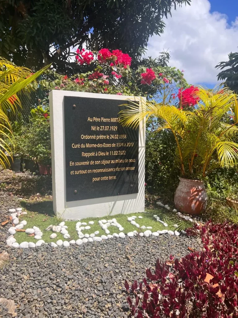 Stèle commémorative en souvenir du Père Pierre Mirta, entourée de plantes.